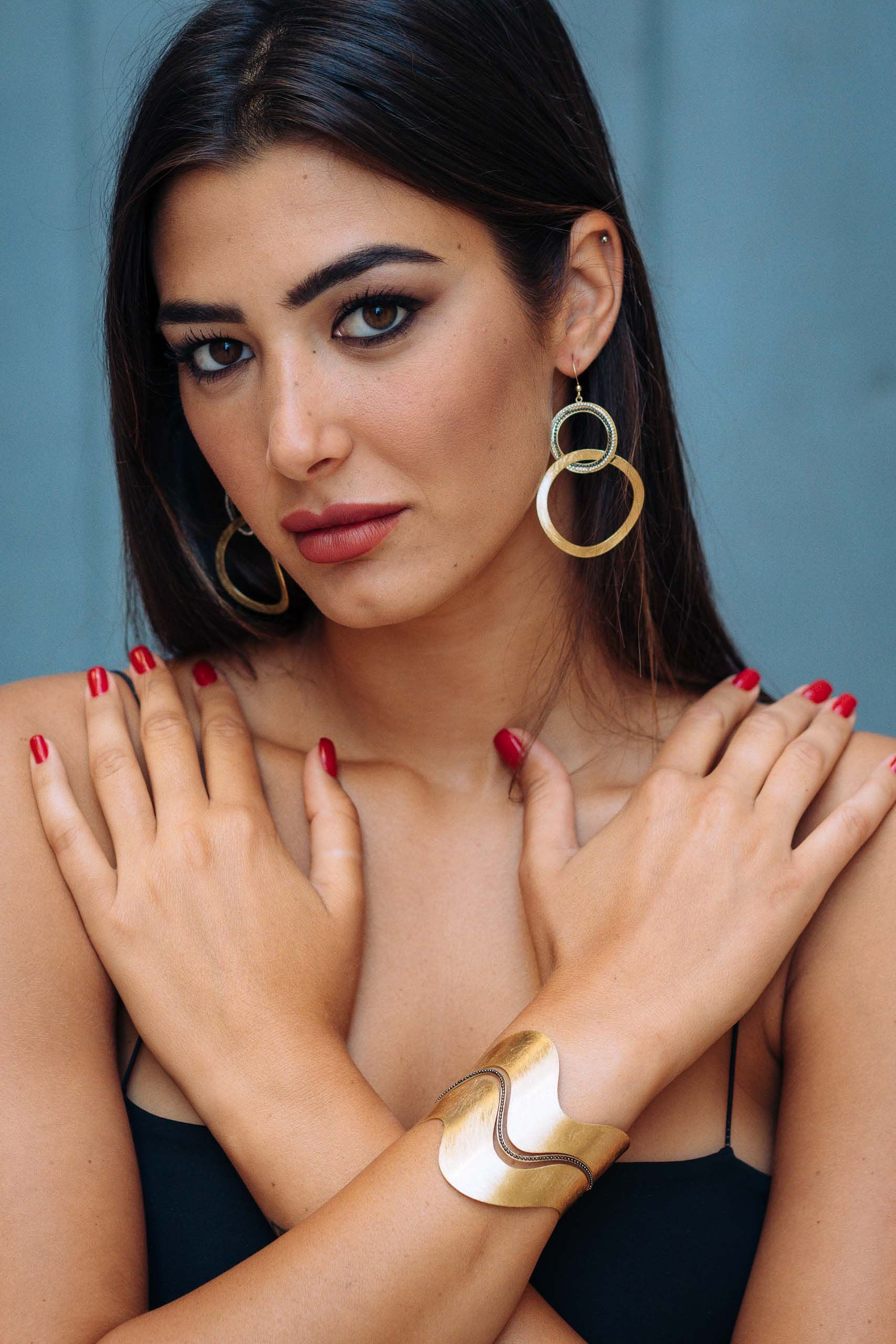 Foto con modella mora di orecchini pendenti graffiati a mano, bagnati in vero oro con componenti in argento chiaro e scuro. Gli orecchini sono formati da due cerchi che si intersecano. Il primo cerchio più vicino al lobo è piccolo con una doppia linea di palline incastonate. Il cerchio sotto è grande e graffiato. Collezione Cleopatra di KLK.